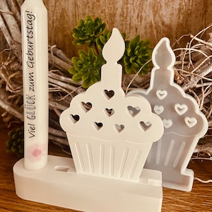 Könnte beinhalten: Weißer Cupcake-Kerzenhalter mit einer einzelnen Kerze und dem Text "Viel Glück zum Geburtstag!" auf der Kerze. Der Halter hat ein Cupcake-Design mit Herzen und einer weißen Basis.