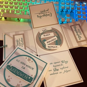 Könnte beinhalten: Eine weiße, graue und türkisfarbene Karte mit einem türkisfarbenen Kreis und dem Text "EAT. SLEEP. ZOCKEN. REPEAT." im Kreis. Die Karte ist zu einer Schachtelform gefaltet. Die anderen Seiten der Schachtel haben den Text "ALLE LIEBEN WÜNSCHE ZUR JUGENDWEIHE", "KLEINER WUNSCHERFALLER", und "ein eigener Weg beginnt da, wo Du aufhörst anderen zu folgen!"