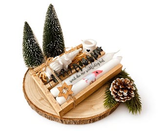 May include: A wooden tray containing two white candles with the German text "weil du mir wicht(el)ig bist" and a gnome illustration, decorated with a wooden star, dried greenery, two miniature pine trees, and a pinecone on a round wood slice.