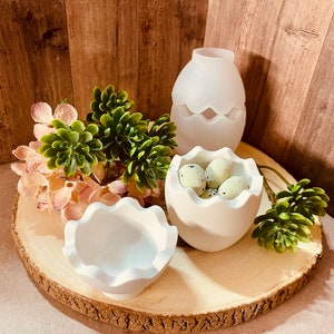 Könnte beinhalten: Drei weiße, eiförmige Behälter aus Keramik, zwei mit gewellten Rändern. Ein Behälter enthält kleine, gesprenkelte grüne Eier. Die Behälter stehen auf einer Holzscheibe mit grünen und rosa Pflanzen.