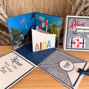 Könnte beinhalten: Eine handgefertigte Pop-up-Grußkarte mit nautischem Motiv, inklusive norwegischer Flagge, Rettungsring und Krabbe. Die Karte zeigt den Text AIDA, Ahoi, Reisegutschein, Norwegen, Auf ins Abenteuer und Die Kunst bedeutet zur richtigen Zeit eine Pause zu machen.