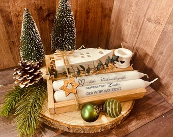 May include: A wooden crate filled with Christmas decorations, including two white candles, a small white house, a candle holder, and eucalyptus sprigs. The crate is sitting on a wooden slice with green and white baubles. The candles have a message that reads "Liebe Weihnachtsgrüße Genies den Zauber der Weihnachtszeit".