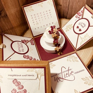 Könnte beinhalten: Eine weiße Hochzeitstorte mit roten und orangen Blumen darauf. Die Torte befindet sich in einer Schachtel mit braunem und rotem Design. Die Schachtel hat auf einer Seite einen Kalender, auf einer anderen Seite die Aufschrift "Gemeinsam ins Glück" und auf einer anderen Seite die Aufschrift "Für Euch". Die Schachtel hat außerdem auf einer Seite die Aufschrift "Magdalene und Denis" und auf einer anderen Seite die Aufschrift "ESUCHT UND GEFUNDEN- in Liebe VERBUNDEN".