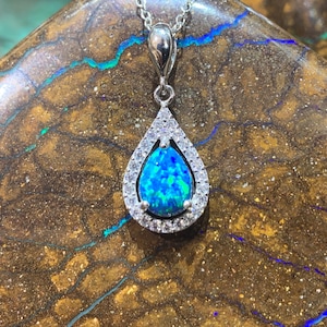 May include: A teardrop-shaped pendant necklace with a blue opal gemstone surrounded by a halo of clear cubic zirconia stones. The pendant is on a silver chain and is resting on a brown and blue patterned surface.