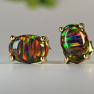 May include: A pair of gold earrings featuring oval-shaped black opal gemstones. The opals display a rainbow iridescent sheen.