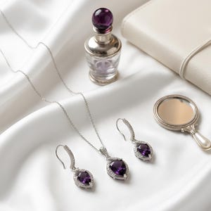 K&ouml;nnte beinhalten: Ein Schmuckset bestehend aus einer silbernen Halskette und zwei passenden Ohrh&auml;ngern, jeweils mit einem birnenf&ouml;rmigen violetten Edelstein, der von kleinen klaren Steinen umgeben ist, neben einem kleinen Glasparf&uuml;mflakon und einem Handspiegel.