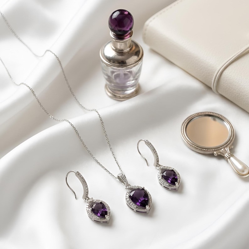 K&ouml;nnte beinhalten: Ein Schmuckset bestehend aus einer silbernen Halskette und zwei passenden Ohrh&auml;ngern, jeweils mit einem birnenf&ouml;rmigen violetten Edelstein, der von kleinen klaren Steinen umgeben ist, neben einem kleinen Glasparf&uuml;mflakon und einem Handspiegel.