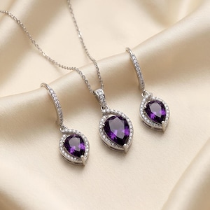 K&ouml;nnte beinhalten: Ein Schmuckset bestehend aus einer Halskette und zwei passenden Ohrh&auml;ngern. Jedes St&uuml;ck zeigt einen birnenf&ouml;rmigen violetten Amethyst, der von einem Kranz aus kleinen klaren Steinen umgeben ist und in silberfarbenes Metall gefasst ist.