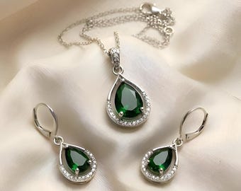 Könnte beinhalten: Ein Silberschmuckset bestehend aus einer Halskette mit Anhänger und zwei passenden Ohrhängern. Jedes Stück zeigt einen birnenförmigen, dunkelgrünen Edelstein, der von einem geschwungenen Silberrand mit kleinen, klaren, weißen Steinen umrahmt ist.