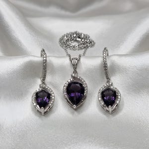 K&ouml;nnte beinhalten: Ein Schmuckset bestehend aus einer Halskette und zwei passenden Ohrringen. Jedes St&uuml;ck zeigt einen birnenf&ouml;rmigen, tiefvioletten Amethyst, der von einem silberfarbenen Rahmen mit kleinen klaren Steinen umgeben ist. Die Kette hat eine feine Gliederkette.