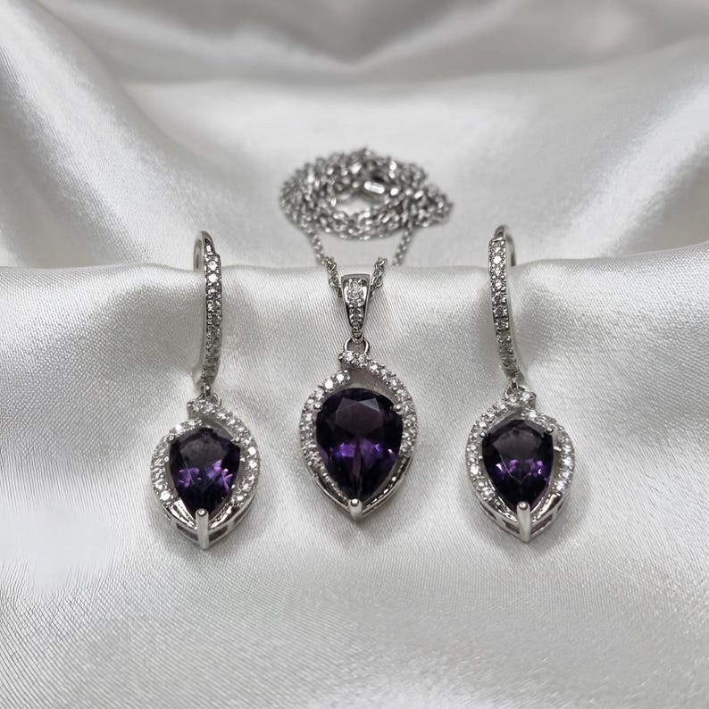 K&ouml;nnte beinhalten: Ein Schmuckset bestehend aus einer Halskette und zwei passenden Ohrringen. Jedes St&uuml;ck zeigt einen birnenf&ouml;rmigen, tiefvioletten Amethyst, der von einem silberfarbenen Rahmen mit kleinen klaren Steinen umgeben ist. Die Kette hat eine feine Gliederkette.