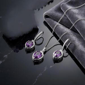 K&ouml;nnte beinhalten: Ein Schmuckset bestehend aus einer silberfarbenen Halskette und zwei passenden Ohrh&auml;ngern. Jedes Schmuckst&uuml;ck zeigt einen birnenf&ouml;rmigen violetten Edelstein, der von einem Kranz aus kleinen klaren Steinen umgeben ist, an einer feinen Kette.