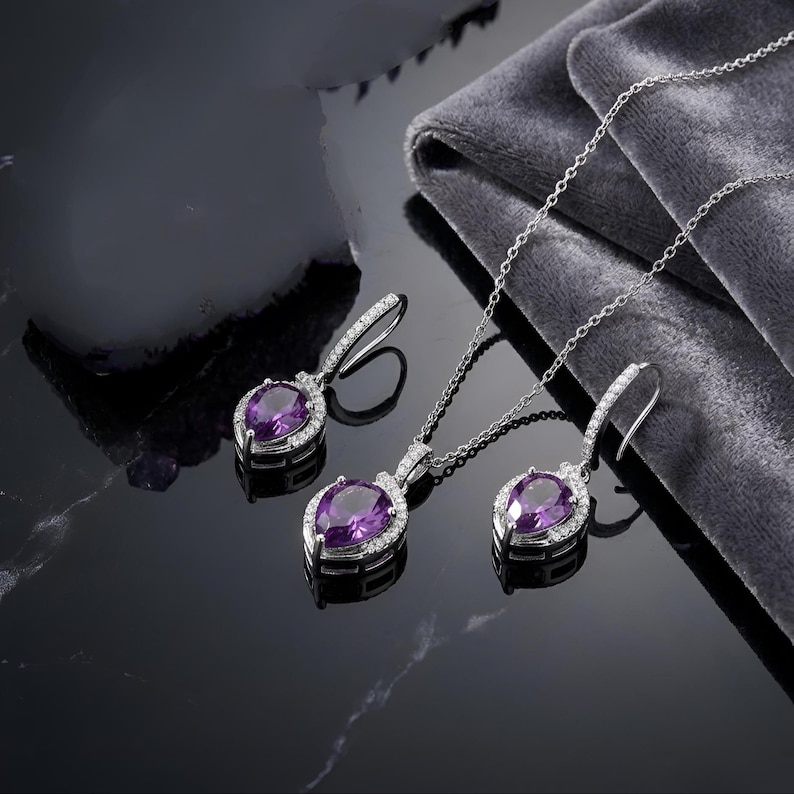 K&ouml;nnte beinhalten: Ein Schmuckset bestehend aus einer silberfarbenen Halskette und zwei passenden Ohrh&auml;ngern. Jedes Schmuckst&uuml;ck zeigt einen birnenf&ouml;rmigen violetten Edelstein, der von einem Kranz aus kleinen klaren Steinen umgeben ist, an einer feinen Kette.