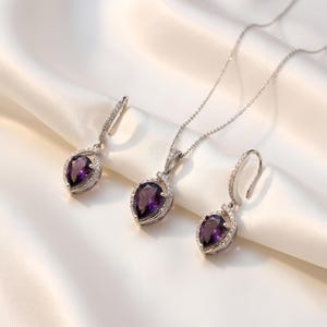 K&ouml;nnte beinhalten: Ein Schmuckset bestehend aus einer Halskette und zwei passenden Ohrringen. Jedes St&uuml;ck zeigt einen birnenf&ouml;rmigen violetten Edelstein, der von einem silberfarbenen Rahmen mit kleinen klaren Steinen umgeben ist. Die Kette hat eine feine silberfarbene Gliederkette.