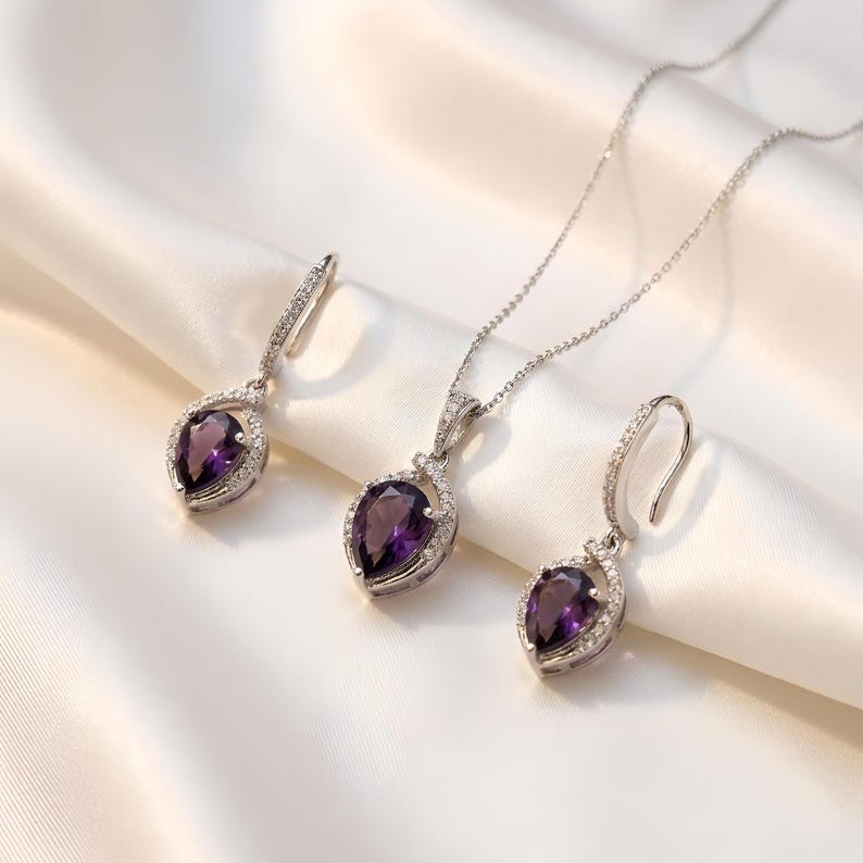 K&ouml;nnte beinhalten: Ein Schmuckset bestehend aus einer Halskette und zwei passenden Ohrringen. Jedes St&uuml;ck zeigt einen birnenf&ouml;rmigen violetten Edelstein, der von einem silberfarbenen Rahmen mit kleinen klaren Steinen umgeben ist. Die Kette hat eine feine silberfarbene Gliederkette.
