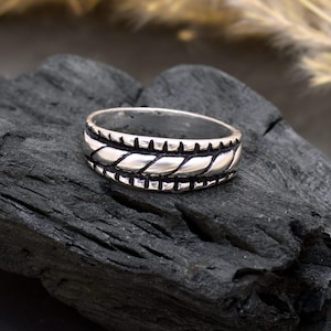 May include: A silver ring featuring a central braided rope design, flanked by rows of small rectangular cutouts. The ring is displayed against a dark, textured backdrop, highlighting its details.
