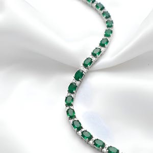 Op de afbeelding: Een zilverkleurige armband met een reeks ovaal geslepen diepgroene stenen in een gebogen lijn. Elke groene steen wordt gescheiden door kleine, heldere, ronde accentstenen die in de metalen schakels zijn gezet, afgewerkt met een sluiting.