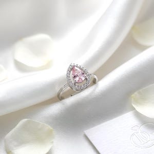 May include: A silver-toned ring featuring a pear-shaped light pink center stone surrounded by a halo of small clear stones. The ring band is thin and metallic, showcasing a delicate design that highlights the central teardrop-shaped gemstone.