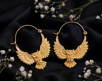 May include: A pair of gold-toned hoop earrings featuring a detailed owl with outstretched wings. The owl design is integrated into the circular hoop structure, showcasing textured feathers and a prominent head, crafted with a matte metallic finish.