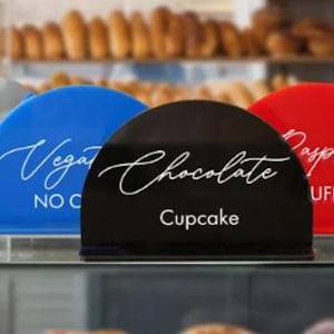 May include: A display of colorful, semi-circular signs with white cursive text. The signs are black, blue, red, gold, and silver. The signs read "Chocolate Cupcake", "Raspberry Muffin", "Blueberry Cookie", "Vegan No Cheese", and "Lemonade Pie".