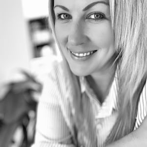 May include: A black and white portrait of a person with shoulder-length hair, wearing a collared shirt with a vertical pinstripe pattern. The person is smiling, looking directly at the camera, with visible eyebrows and a collared neckline.