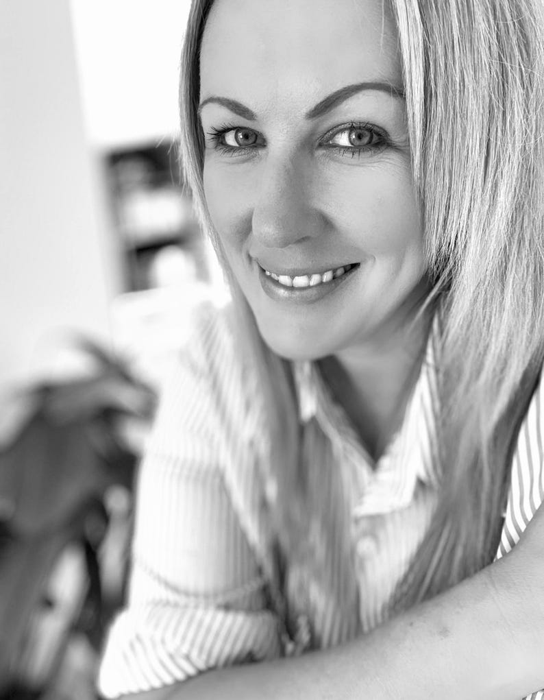 May include: A black and white portrait of a person with shoulder-length hair, wearing a collared shirt with a vertical pinstripe pattern. The person is smiling, looking directly at the camera, with visible eyebrows and a collared neckline.