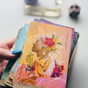 May include: A tarot card titled Queen of Wands featuring a profile illustration of a person with a shaved head adorned with red and yellow flowers, wearing a yellow and orange garment, set against a warm gradient background with pink fluid patterns.