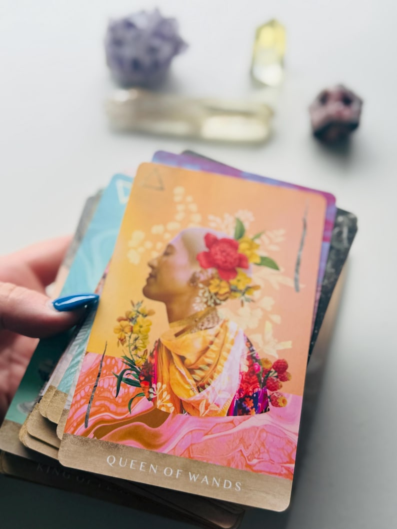 May include: A tarot card titled Queen of Wands featuring a profile illustration of a person with a shaved head adorned with red and yellow flowers, wearing a yellow and orange garment, set against a warm gradient background with pink fluid patterns.