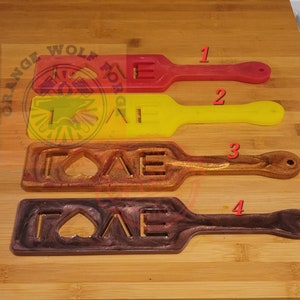 May include: Four paddle-shaped objects in red, yellow, and two shades of brown. The red and yellow paddles have the letter "E" cut out. The brown paddles have the word "LOVE" cut out. The objects are on a wooden surface.