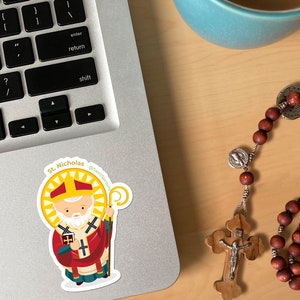 May include: A sticker of Saint Nicholas, a cartoon illustration of a man with a white beard wearing a red and white bishop's robe, holding a book and a staff. The sticker is on a silver laptop with a keyboard and a blue coffee mug in the background.