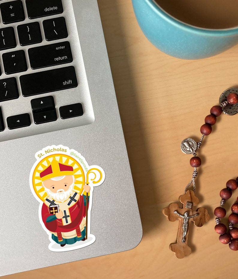 May include: A sticker of Saint Nicholas, a cartoon illustration of a man with a white beard wearing a red and white bishop's robe, holding a book and a staff. The sticker is on a silver laptop with a keyboard and a blue coffee mug in the background.