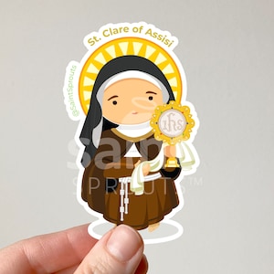 May include: A sticker featuring a cartoon illustration of Saint Clare of Assisi, a Catholic saint. She is depicted in a brown habit with a white veil and holding a monstrance with the letters "IHS" on it. The sticker has a yellow sunburst behind her and the text "St. Clare of Assisi" at the top.