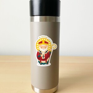 May include: A stainless steel water bottle with a sticker of Saint Nicholas, a cartoon illustration of a man with a white beard wearing a red and gold bishop's robe, holding a staff and a book. The sticker has the text "St. Nicholas" and "@saint.scott".