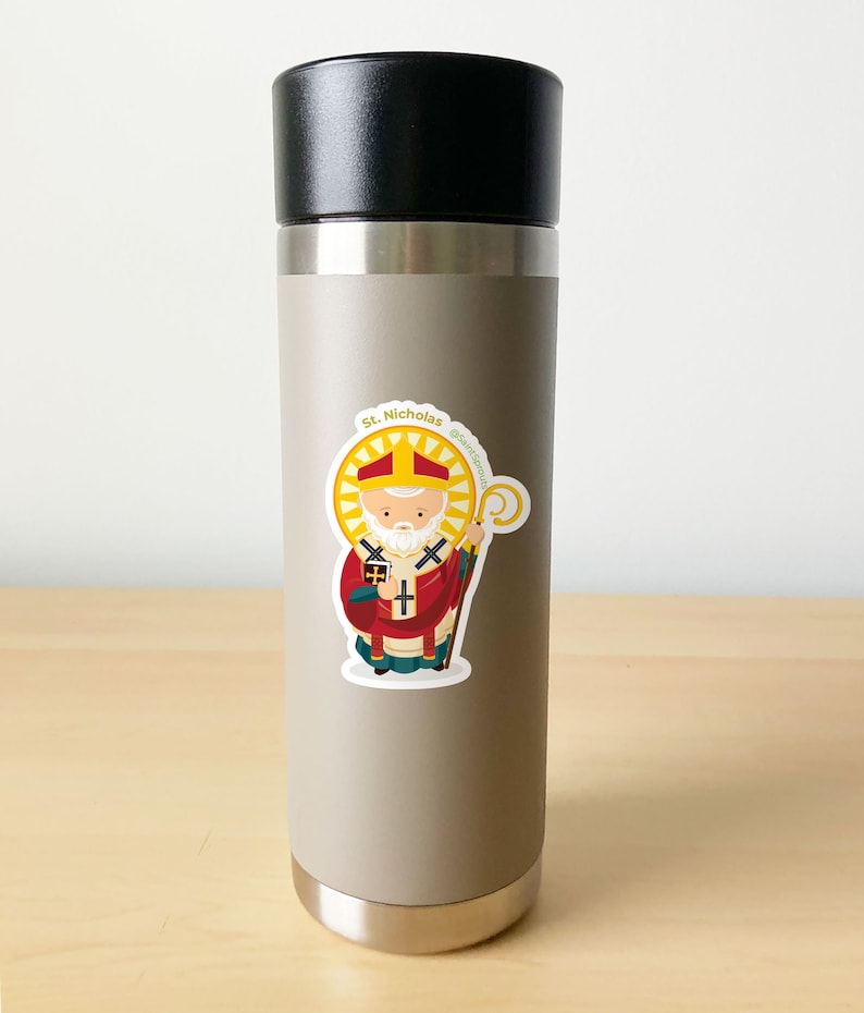 May include: A stainless steel water bottle with a sticker of Saint Nicholas, a cartoon illustration of a man with a white beard wearing a red and gold bishop's robe, holding a staff and a book. The sticker has the text "St. Nicholas" and "@saint.scott".