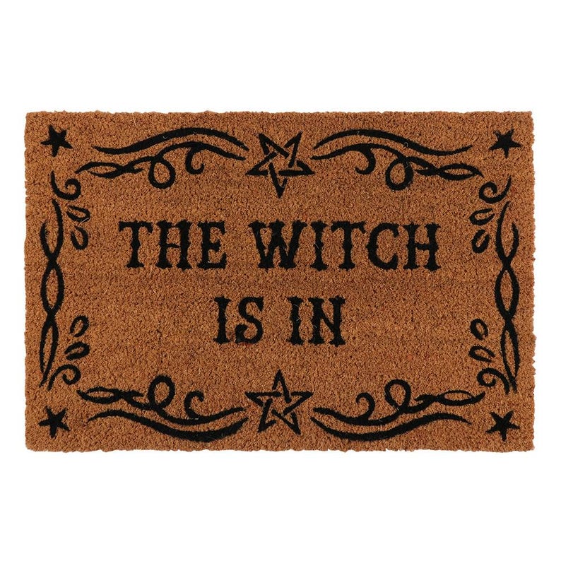 May include: Brown rectangular doormat with the text "THE WITCH IS IN" in black. The text is surrounded by a black decorative border with stars and swirls. The doormat is made of a coarse, natural fiber material.