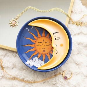 May include: A blue trinket dish with a sun and moon design. The sun is orange, and the moon is yellow. Gold necklaces with sun and moon pendants are also visible. The dish is likely ceramic and suitable for small items.