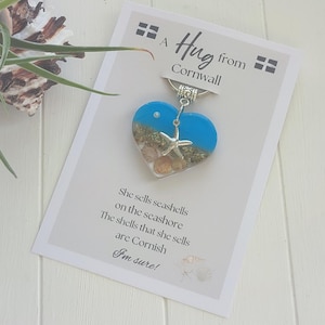 May include: A heart-shaped pendant with a blue and sand design, featuring a starfish charm. The pendant is attached to a card that reads "A Hug from Cornwall" and a poem about seashells.