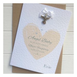 May include: A white card with the words "Angel Baby" and the text "In Heaven Above, Never Forgotten, Forever Loved" inside a peach heart. A silver angel charm and white bow are attached to the top. The name "Evie" is printed at the bottom.