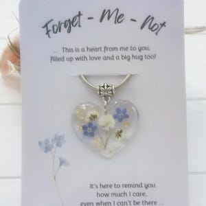 May include: A heart-shaped resin keyring containing pressed blue and white forget-me-not flowers and gold flakes. It is attached to a silver-toned metal ring and presented on a white card printed with the text Forget-Me-Not and sentimental messages.