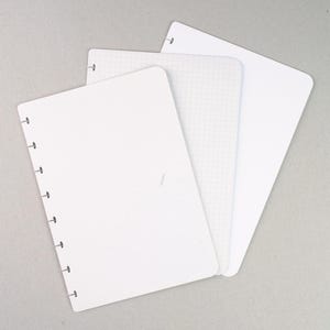 Może przedstawiać: Trzy białe strony notatnika z oprawą dyskową. Jedna strona jest pusta, druga ma wzór kratki, a trzecia również jest pusta. Każda strona ma otwory wzdłuż grzbietu do oprawy. Strony są ułożone na jasnoszarej powierzchni.