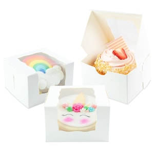 May include: Three white bakery boxes with clear windows showcasing decorated treats. One box displays a rainbow cake, another a cupcake with pink frosting and a strawberry, and the third a unicorn-themed cookie.