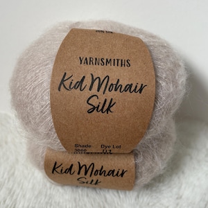 Puede incluir: Una madeja de hilo Yarnsmiths Kid Mohair Silk en color beige claro. La etiqueta del hilo es marrón con texto negro que dice "Kid Mohair Silk". La etiqueta también indica el tono y el lote de tinte.