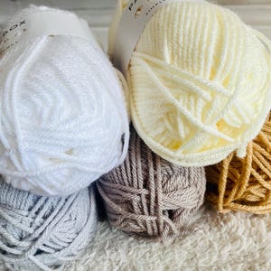 May include: Close-up of several skeins of chunky yarn in various colors. The colors include white, light yellow, gray, taupe, and a golden brown. The yarn is tightly wound and appears soft, ideal for knitting or crocheting projects.