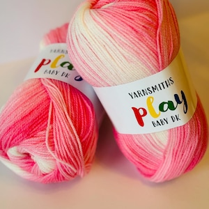 Könnte beinhalten: Zwei Knäuel rosa und weißes Garn mit dem Etikett "Yarnsmiths Play Baby DK" auf einem Knäuel.