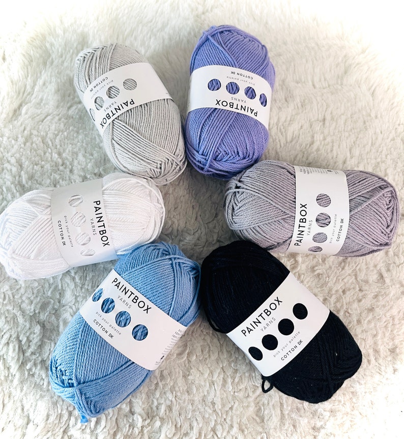 May include: Six balls of yarn in various colors, including white, light blue, dark blue, gray, and purple. Each ball has a label that says "Paintbox Yarns" and includes the text "Cotton DK".