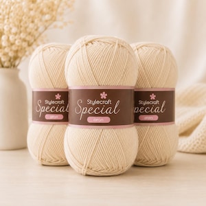 May include: Three beige balls of Stylecraft Special aran yarn, each with a brown and pink label. The yarn is tightly wound and appears soft. The image is a close-up shot, with a vase of flowers and a folded blanket in the background. The yarn is suitable for knitting.