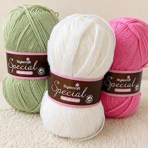 Könnte beinhalten: Drei Knäuel Stylecraft Special Doppelstrickgarn in Grün, Weiß und Pink. Jedes Knäuel hat ein braunes Etikett mit dem Markennamen und einem rosa Rand. Ideal für Strick- und Häkelprojekte.