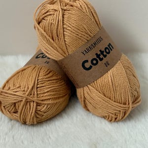 May include: Two skeins of Yarnsmiths Cotton DK yarn in a warm, golden-brown colour. Each skein is wrapped with a brown paper label. The yarn is tightly wound, perfect for knitting or crochet projects.