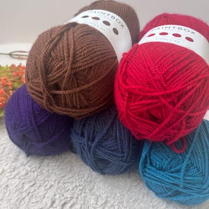 Puede incluir: Cinco ovillos de lana en diferentes colores: marrón, rojo, morado, azul y verde azulado. Cada ovillo tiene una etiqueta blanca con el texto "Paintbox Simply DK".