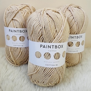以下が含まれることがあります： ベージュ色の糸玉が3つ。白いラベルには「PAINTBOX YARNS」と「COTTON DK」と書かれています。糸はきつく巻かれており、ラベルには一連の円があります。糸は柔らかい白い表面に展示されています。
